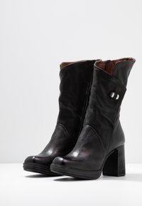 Ett par svarta läderankelstövlar med klack, sidogömma och dekorativt silverspänne i detalj mot en vit bakgrund.