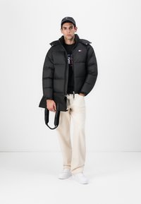 Svart pufferjakke med glidelås og hette. Modellen har på seg en svart t-skjorte og lyse beige bukser, og holder en svart totebag med tekst.