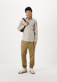 Gaisms kvadrātains apakšveļas krekls bēšā un brūnā krāsā, khaki šaurlās bikses un baltas kedas ar melniem akcentiem; tur rokassomu melnā krāsā.