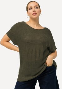 Olivgrünes Stricktop mit lockerem Schnitt, weitem Ausschnitt und kurzen Ärmeln. Verfügt über ein strukturiertes Design mit einem zentralen Motiv. Kombiniert mit dunkelblauen Jeans.