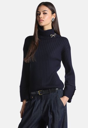 Femme aux longs cheveux foncés portant un pull à col roulé côtelé noir, un pantalon taille haute sombre et une broche sur le col.