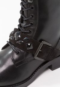 Botte noire en cuir à lacets avec une sangle décorative en daim ornée de pierres précieuses noires taillées et d'une boucle en métal.