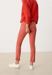 Lichtroze spijkerbroek met een slim fit en opgerolde boorden, gecombineerd met een oranje gebreid trui en witte sneakers. Egaal achtergrond.