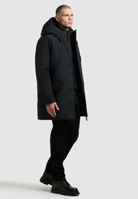 Czarna parka z dużym kapturem i zapięciem na zamek z przodu, z bocznymi kieszeniami i elegancką, gładką fakturą. Noszona na czarne spodnie i buty.