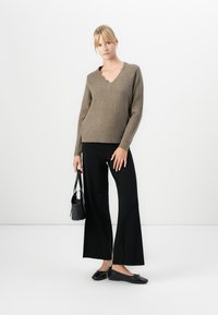 Femeie stând în fața unui fundal alb, îmbrăcată într-un pulover maro cu decolteu în V, pantaloni negri largi, balerini negri și ținând o geantă negre.