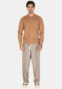 Maglione marrone chiaro con scollatura a girocollo, abbinato a pantaloni grigi larghi con coulisse. Il modello è in piedi su uno sfondo semplice.