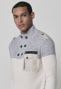 Suéter de punto con un alto cuello, rayas en gris y crema, bolsillo con cremallera en la parte frontal y detalles en botones. Tejido texturizado con patrón acanalado.