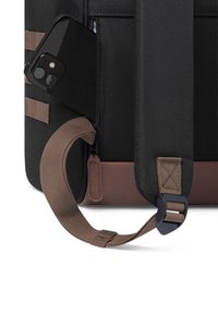 Zaino nero con dettagli marroni, dotato di una tasca laterale e di uno smartphone parzialmente visibile. Tessuto testurizzato e dettagli della tracolla regolabile.