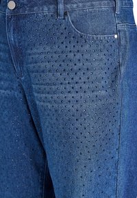 Jeans de mezclilla ajustados con un lavado azul oscuro. En la parte delantera tienen un patrón de pequeños tachuelas negras reflectantes dispuestas en filas.