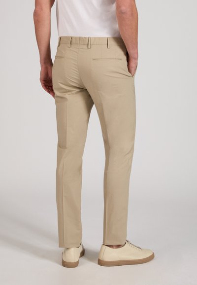 Homme portant un pantalon beige slim-fit et des baskets blanc cassé, debout les mains dans les poches, vu de dos sur un fond uni.