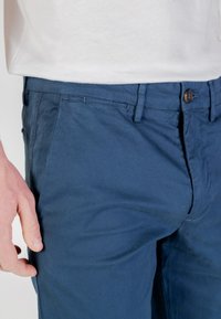 Chino in cotone blu navy con una consistenza liscia, dotato di chiusura a bottone e tasche laterali. Le pieghe visibili esaltano i dettagli del tessuto.