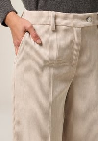 Pantalones de pana beige con estrías verticales, bolsillo frontal y cierre de botón de metal en la cintura. Tejido suave y texturizado.