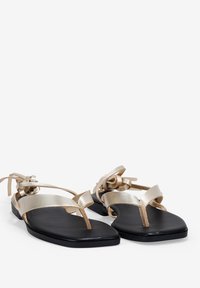 Sandalias planas con una base negra y tiras metálicas doradas. Presentan un diseño tipo thong con acentos en torsión y un acabado de cuero suave.