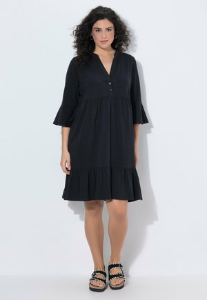 Kobieta o kręconych włosach, ubrana w luźną czarną sukienkę z rękawami 3/4 i falbanami u dołu, zestawioną z czarnymi sandałami z białymi detalami.