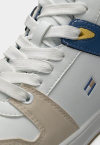 Λευκό δερμάτινο αθλητικό παπούτσι με μπεζ λεπτομέρειες, μπλε λουράκι και κίτρινα στοιχεία. Τα κορδόνια και οι τρύπες είναι εμφαντικά ορατά. Διακριτή υφή.