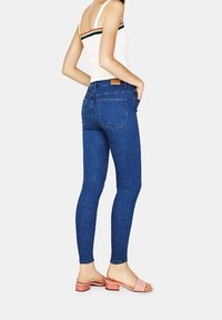 Hubené modré džínové kalhoty s polohou v pase, zadní kapsy a texturovaným povrchem, v kombinaci s bílým topem na ramínka a růžovými pantoflemi.