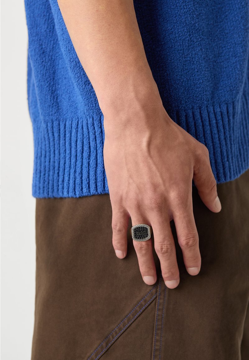 Pulover tricotat texturat albastru cu tiv ondulat, purtat cu pantaloni din țesătură maro; o mână ține un inel dreptunghiular negru și argintiu cu detalii intricate.
