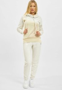 Hellbeige Kapuzenpullover mit Schalkragen und Kordeln, kombiniert mit passenden Jogginghosen. Weicher Stoff, dezente Textur und gesprenkelte Designdetails.