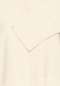 Kremės spalvos megztinis pagamintas iš reljefo trikotažo, su V formos iškirpte, ilgomis rankovėmis ir penkiais plastikiais mygtukais priekyje.