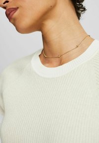 Goldene Halskette mit kleinen, abgerundeten Akzenten, kombiniert mit einem cremefarbenen, gerippten Pullover mit Rundhalsausschnitt und strukturiertem Stoff.