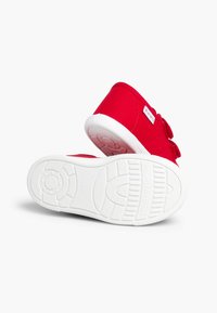 Zapatillas de lona rojas con suela de goma blanca, punta redondeada y correa de Velcro para un fácil ajuste. Diseño sencillo con un detalle de marca sutil.