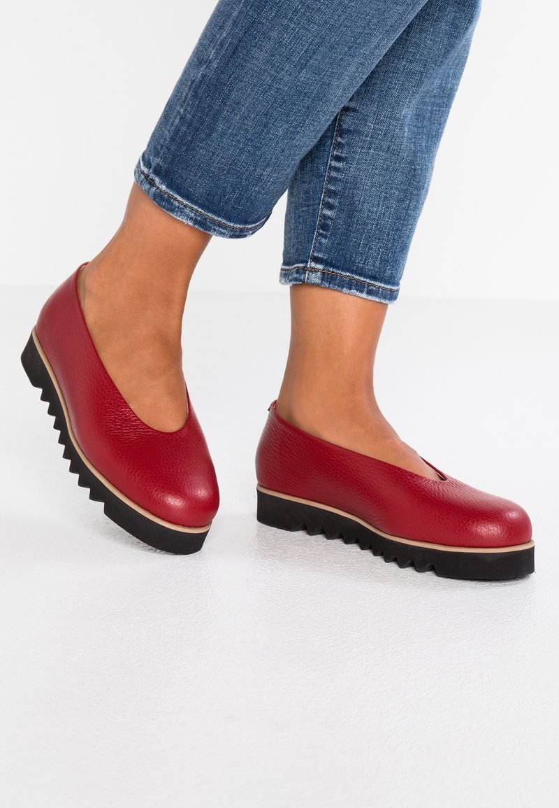 Pieds portant des chaussures rouges en cuir sans lacets avec des semelles épaisses noires rainurées, associées à un jean bleu retroussé, debout sur un sol blanc.
