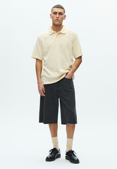 Világos sárga texturált galléros póló, fekete farmer rövidnadrág, krémszínű zokni és fekete cipő. A pólón gallér és rövid ujj található.