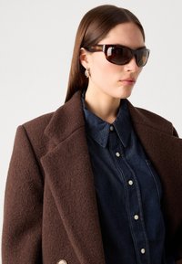 Cappotto di lana marrone con un colletto strutturato, abbinato a una camicia di denim con chiusura a bottoni. Occhiali da sole marroni con una tonalità scura.