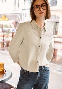 Jeune personne portant de grandes lunettes brunes, une chemise beige boutonnée et un jean, debout à l'intérieur près d'une fenêtre en verre avec un café à l'extérieur.