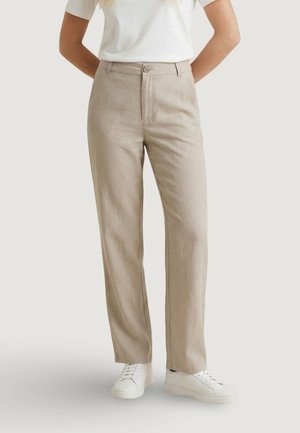 Pantaloni beige a gamba dritta indossati con scarpe da ginnastica bianche e una maglietta bianca a maniche corte, persona in piedi con le mani dietro la schiena.