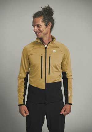 Mann trägt eine beige-schwarze, durchgehende Jacke mit zwei vertikalen Brusttaschen, steht vor einem schlichten grauen Hintergrund, schaut nach unten und lächelt.