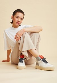 Sneaker alte bianche con dettagli grigi e colletto rosso, dotate di suola testurizzata e lacci neri. Modello che indossa pantaloni beige a gamba larga e una maglietta bianca.