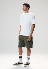 Maglietta bianca in cotone a maniche corte, pantaloncini cargo verdi, sneakers nere e calzini a righe con accenti verdi e blu navy. Modello in piedi di lato.