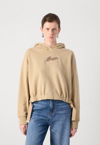 Beige avkortet hettegenser med snor i bunnen og brodert logo. Har lange ermer og myk stoff. Kombinert med blå jeans.