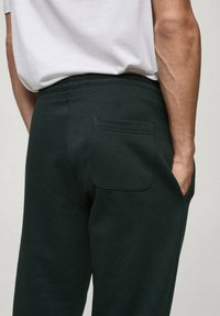 Temnozeleni trenirke iz mehkega blaga, ki imajo zadnji žep, elastični pas in sproščen kroj. Minimalističen dizajn in čiste linije.