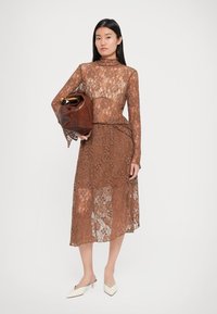 Femme portant une robe en dentelle florale marron, tenant un grand sac à main en cuir marron, assortie de chaussures à talons bas pointues blanches, se tenant sur un fond blanc.