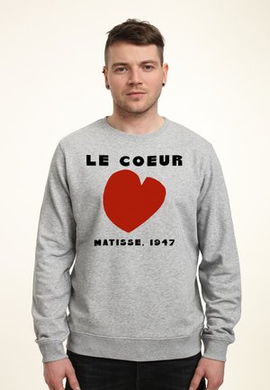 Mężczyzna w szarej bluzie z dużym czerwonym sercem i napisem "Le Coeur Matisse 1947" z przodu, stojący na jednolitym jasnym tle.