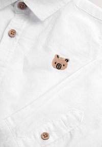 Camicia bianca con colletto, due bottoni marroni e logo ricamato di un orso in marrone chiaro e nero sulla parte sinistra del petto.