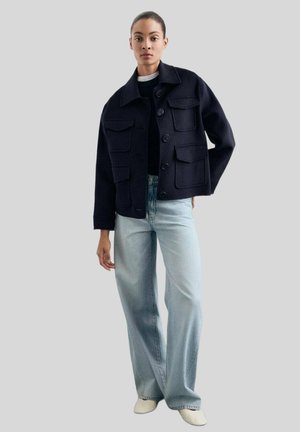 Woman wearing a dark navy buttoned jacket with large pockets, light blue wide-leg jeans, and white shoes against a plain light background.