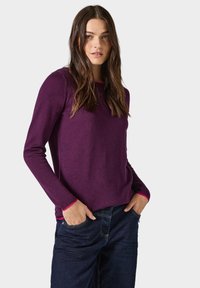 Lila gestrickter Pullover mit rundem Halsausschnitt, der kontrastierende pinke Akzente an den Bündchen aufweist. Getragen mit dunklen Denim-Jeans.