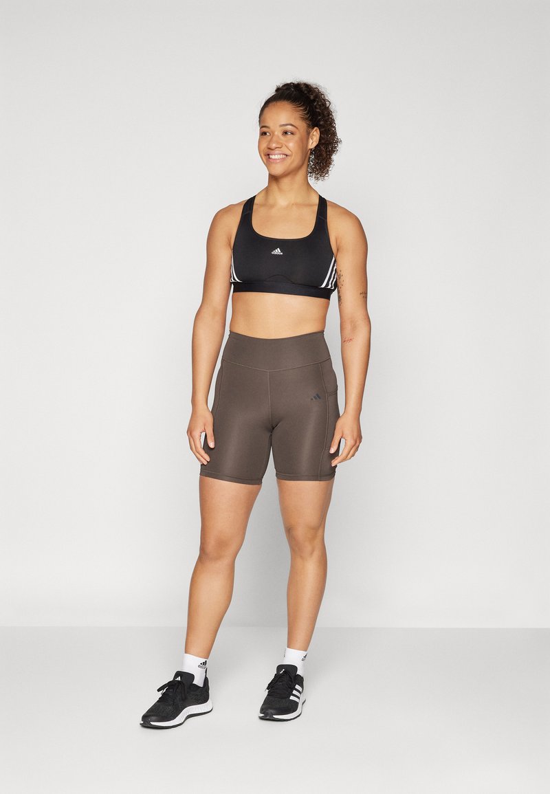 Sujetador deportivo negro con rayas blancas, pantalones cortos marrones ajustados y zapatillas deportivas negras. Tejido suave y cintura alta para mayor comodidad.