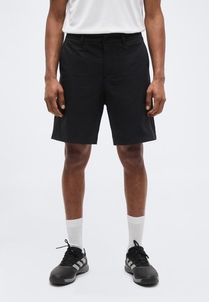 Άνδρας που φοράει μαύρα σορτς μέχρι το γόνατο, λευκές κάλτσες, μαύρα αθλητικά παπούτσια και λευκό πουκάμισο, στέκεται μπροστά σε απλό φόντο.