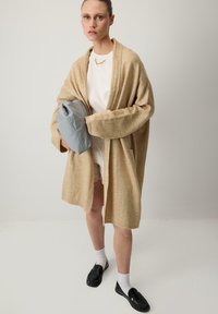 Beżowy sweter dzianinowy z długimi rękawami, luźny krój i otwarty przód. W zestawie z białą bluzką, jasnymi szortami i szarym torbą. Na nogach czarne mokasyny.