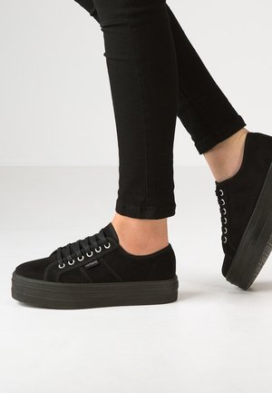 Personne portant des baskets plateformes noires et un pantalon noir ajusté, debout sur un sol clair avec un talon levé.