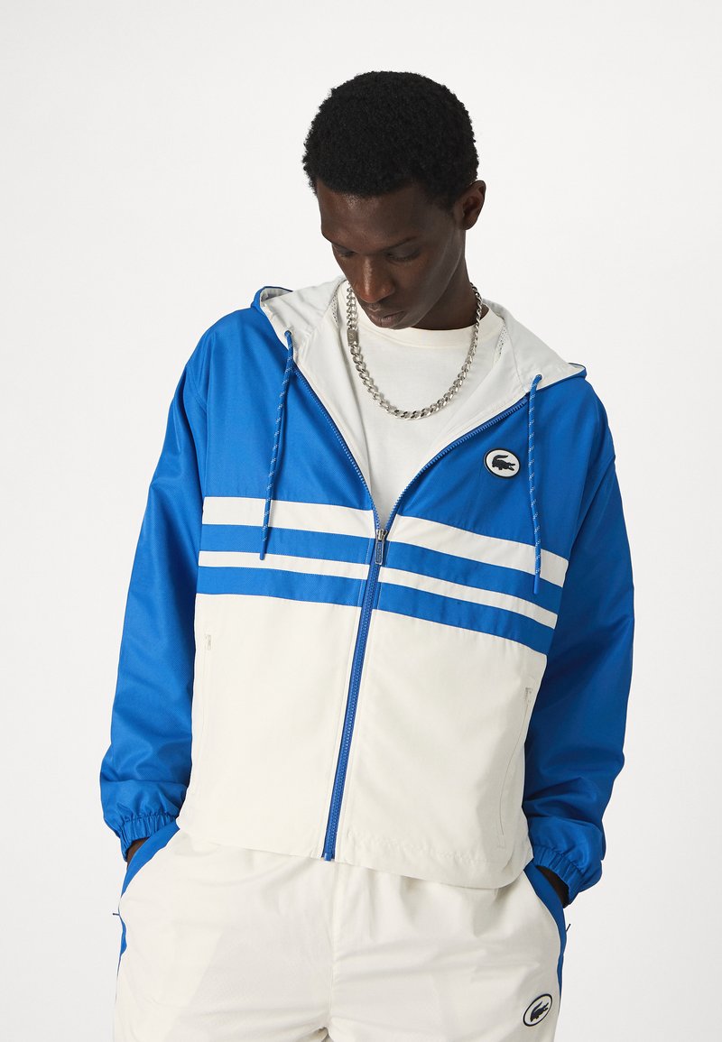 Giacca a vento blu e bianca, con zip frontale, cappuccio regolabile, polsini elastici e design bicolore con strisce e dettagli del logo.