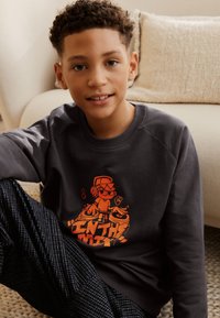 Dunkelgrauer Pullover mit einer orangefarbenen Grafik eines DJ-Charakters. Schwarze und weiße gemusterte Jogginghosen sichtbar, sitzend auf einer gewebten Matte.