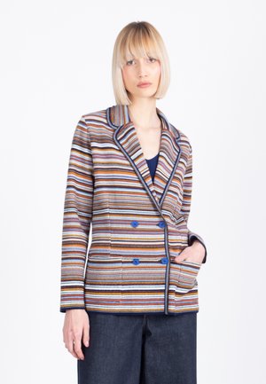 Kobieta o krótkich blond włosach, ubrana w kolorową, wielopasmową, dwurzędową marynarkę i ciemne spodnie, stojąca z jedną ręką w kieszeni.