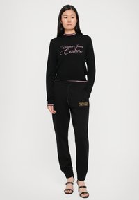Svart langermet genser med rosa "Versace Jeans Couture" logo og matchende svarte sweatpants med gull logo, brukt med svarte spissede sandaler.