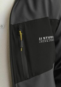Zwart en grijs zip-up jack met een ritszak en een gele trekkoord, en witte logotekst op de borst. Soepele textuur.