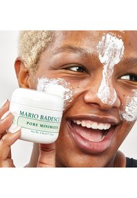 Weißes Glas mit der Aufschrift "Mario Badescu Pore Minimizer", das neben einem Gesicht mit aufgetragenener Tonmaske gehalten wird, was eine glatte, leicht strukturierte Oberfläche betont.